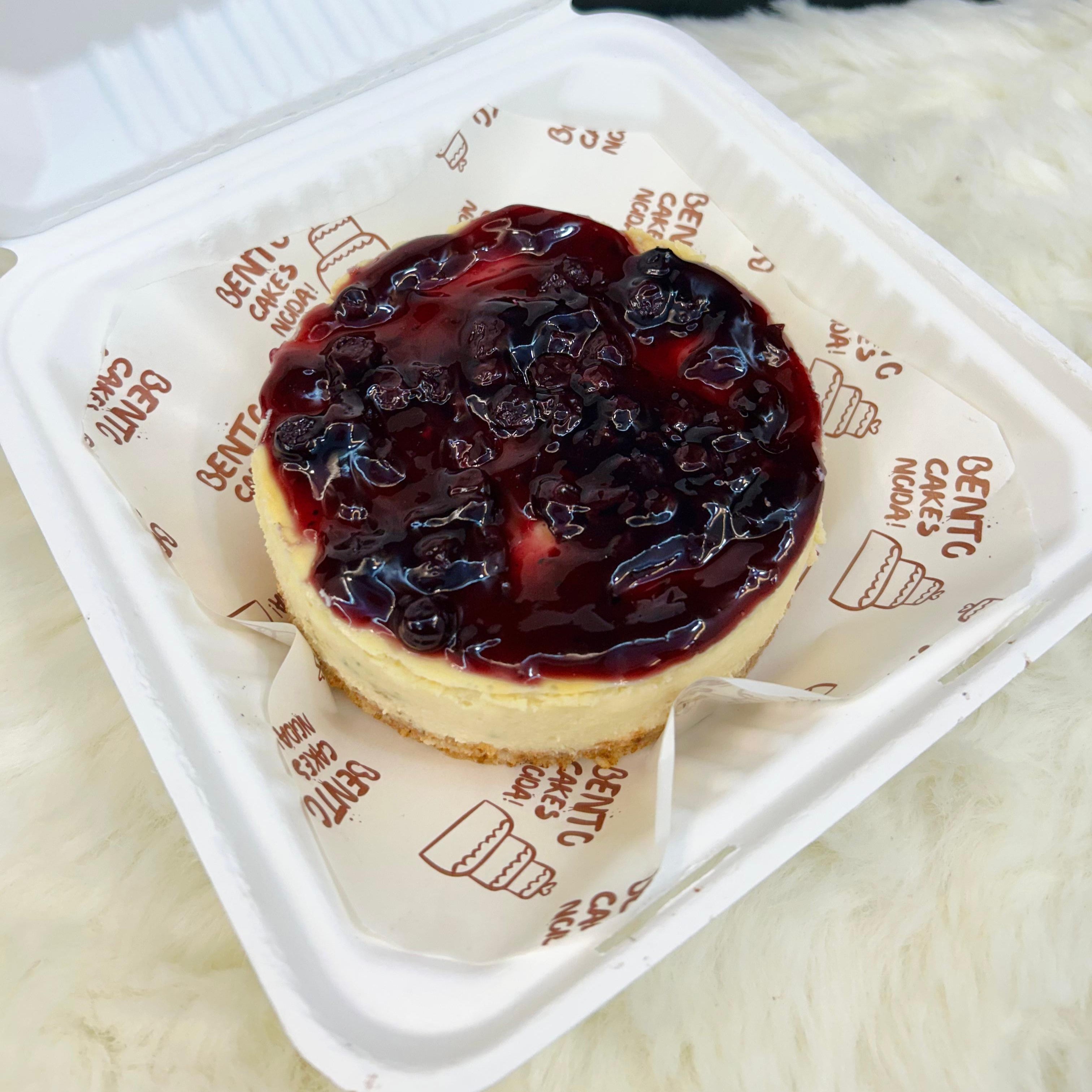 A rich and creamy blueberry cheesecake presented in a white bento-style container, topped with a glossy layer of blueberry compote and garnished with fresh blueberries, set against a minimalistic background with front view.
